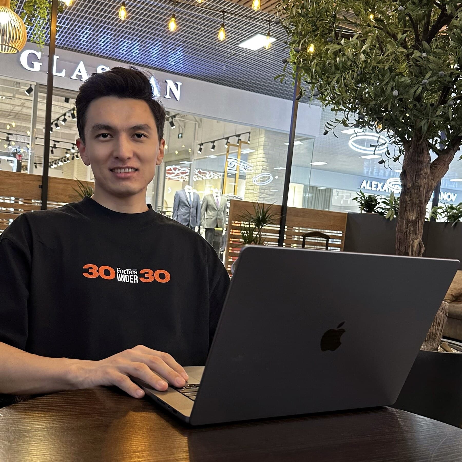 Yerzhan Karatayev in a Forbes 30 Under 30 shirt working on a laptop